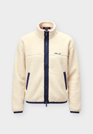 Persona que lleva una chaqueta de forro polar color crema con cremallera y bolsillos negros, pantalones negros, calcetines blancos y zapatillas negras con cordones rojos, de pie quieta.