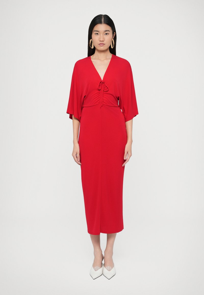 Sarkana midi kleita ar īsām piedurknēm, jostas mezgls un gluda auduma faktūra. Kombinēta ar baltām kurpēm ar noapaļotu purngalu un lielām lokveida auskaru riņķiem.