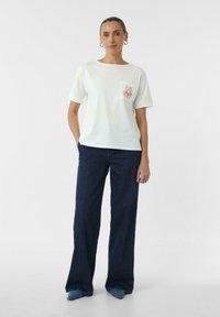 T-shirt en coton blanc avec un imprimé de poche rouge, col rond, manches courtes, accompagné de jeans évasés bleu marine et de chaussures pointues bleues.