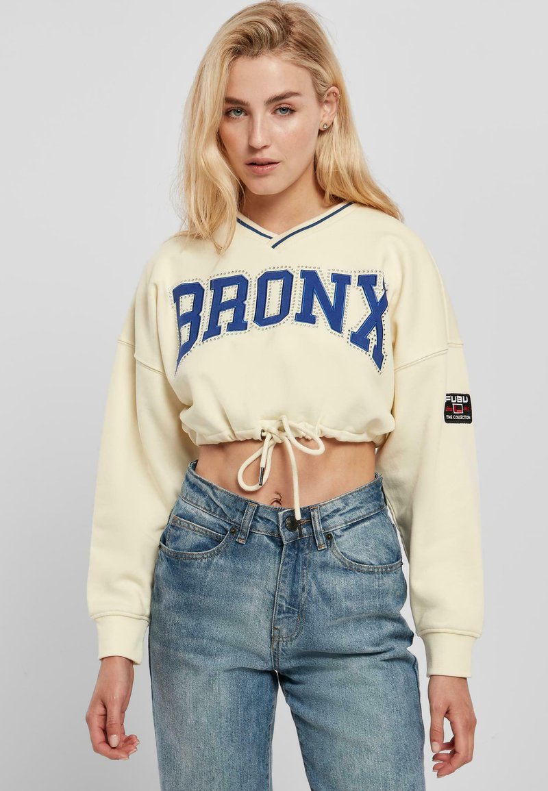 Cropped cream sweatshirt with "BRONX" in blue, drawstring hem, and v-neck; paired with light blue high-waisted jeans.