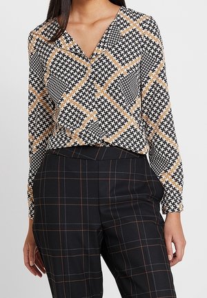 Femme portant un chemisier à manches longues à motifs avec des formes géométriques noires, blanches et beiges et un pantalon à carreaux noir avec de fines lignes marron.