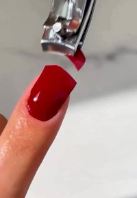 Un tagliaunghie che rifila un'unghia lucida di un profondo rosso con punta squadrata. L'unghia ha una texture liscia, mostrando i dettagli della brillantezza dello smalto.