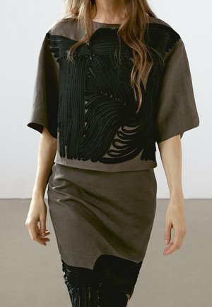 Kobieta w sukience w kolorze taupe i luźnej czarnej bluzce z teksturowanymi, abstrakcyjnymi czarnymi zdobieniami na przodzie i panelu spódnicy.