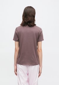 Άτομο με καστανά μαλλιά μέχρι τους ώμους στέκεται στραμμένο προς τα πίσω, φορώντας ένα κοντομάνικο μαβί μπλουζάκι και ανοιχτό ροζ παντελόνι, μπροστά σε λευκό φόντο.