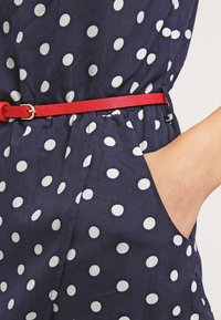 Navy blue fabric with white polka dots, featuring a red leather belt and side pockets. The material has a smooth texture.