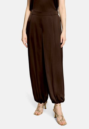 Frau trägt locker sitzende dunkelbraune Hose mit elastischen Bündchen, kombiniert mit goldfarbenen, Riemchen-High-Heel-Sandalen vor einfarbigem Hintergrund.