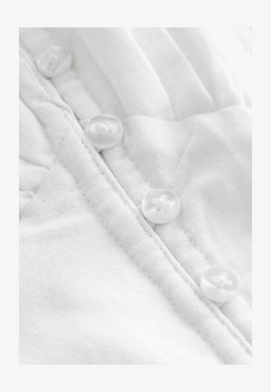 Tissu blanc avec une texture subtile, mettant en avant quatre boutons transparents alignés verticalement le long de la couture. Détails de couture visibles près des boutons.