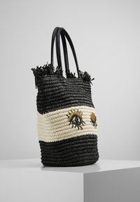 Sac fourre-tout tissé en noir et crème avec un haut à franges. Présente des yeux brodés en or et noir. Poignées longues noires. Finition texturée.
