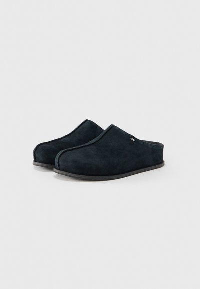 Paire de chaussons en daim noir à enfiler, à bout fermé et petit talon, sur fond blanc.
