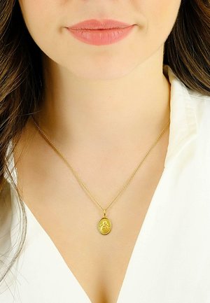 Collier en pendentif en or avec un charme ovale présentant une figure détaillée, suspendu à une chaîne délicate, porté sur une blouse blanche.