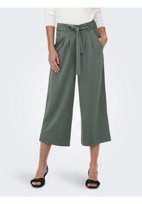 Zelené kalhoty širokého střihu s páskem v pase, lištami vpředu a hladkým povrchem. Nosené s černými sandály na řemínky.