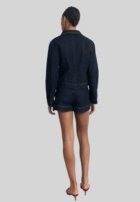 Ensemble en denim foncé comprenant une veste et un short, avec des coutures contrastantes, une coupe ajustée et une silhouette cropped. Porté avec des sandales à talons.