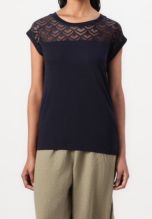 Woman wearing navy blue short-sleeve top with sheer patterned upper chest panel and light beige loose pants, standing against white background.