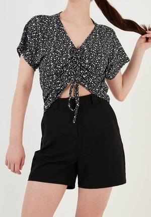 Femme portant un crop top froncé à manches courtes noir à motifs floraux et un short noir taille haute, tenant sa longue queue de cheval d'une main.