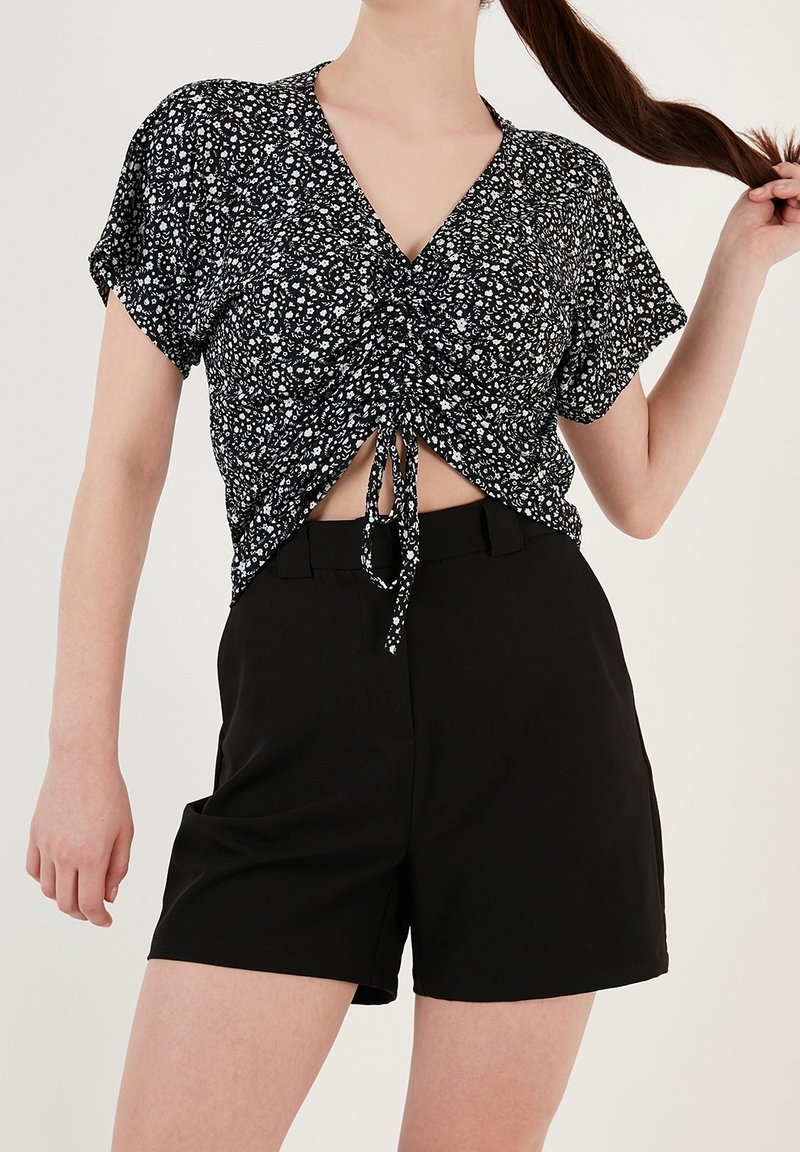 Femme portant un crop top froncé à manches courtes noir à motifs floraux et un short noir taille haute, tenant sa longue queue de cheval d'une main.