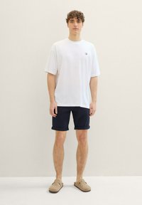 Biała koszulka z krótkim rękawem z małym logo, zestawiona z podwiniętymi granatowymi szortami i beżowymi butami wsuwanymi, na neutralnym tle.