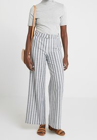 Breda randiga byxor i vitt, marinblått och ljusgrått, med en bälteshög midja, kombinerade med en grå kortärmad topp och bruna sandaler.