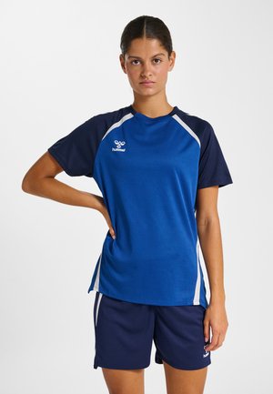 Junge Frau steht mit der Hand in der Hüfte, trägt blau-navyfarbene Sportbekleidung mit weißen Akzenten vor einem schlichten weißen Hintergrund.