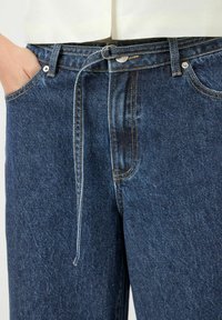 Modré džínové kalhoty s vysokým pasem, poutky na pásek a jedním vázacím detailem. Jsou viditelné výrazné švy a boční kapsy.