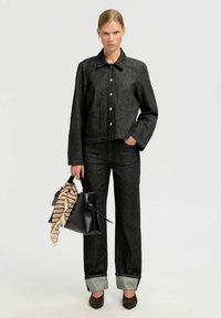 Black denim jacket with silver buttons and two front pockets, paired with matching black denim pants with turned-up gray cuffs. Holding a black bag with a scarf.