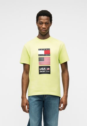 Άνδρας που φοράει ανοιχτό πράσινο T-shirt Tommy Hilfiger με τη σημαία των ΗΠΑ και κείμενο, συνδυασμένο με μπλε τζιν, στέκεται μπροστά σε απλό φόντο.