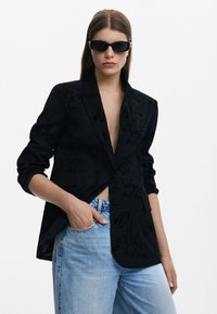 Svart blazer med en subtil texturdesign, enkelknäppning, slag med knappproppar och något lös passform, kombinerad med ljusblå jeans.