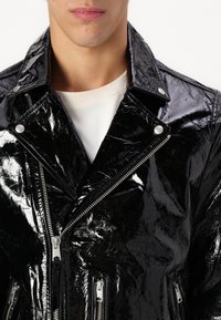Sort, skinnende bikerjakke lavet af blankt materiale med lynlåslukning foran, trykknapper og flere lynlåslommer. Teksturert finish synlig.