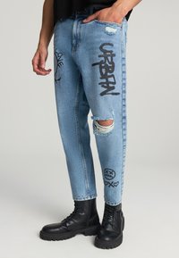 Jeans in denim azzurro con dettagli strappati, testo in stile graffiti nero e accenti grafici, abbinati a stivali da combattimento neri.