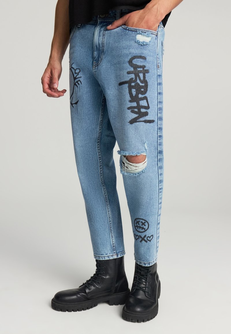 Jeans in denim azzurro con dettagli strappati, testo in stile graffiti nero e accenti grafici, abbinati a stivali da combattimento neri.