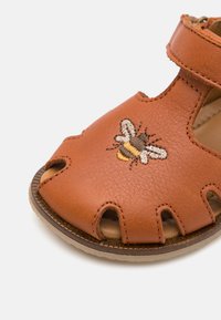 Brun lädersandal med utskärningsdesign och rem. Har en broderad bie-detail i svart, gult och vitt. Platt sula.
