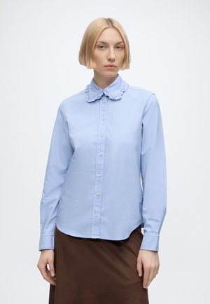 Vrouw met kort blond haar die een lichtblauw blouseje met knopen en een gerimpelde kraag en een bruine rok draagt tegen een effen lichte achtergrond.