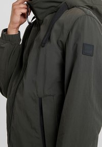 Dark green hooded jacket features textured fabric, front zipper, elastic cuffs, and a square black logo patch on the sleeve.