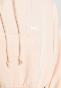 Sweat-shirt de couleur pêche avec une poche avant, des cordons de serrage et un logo "levi's" blanc. Tissu doux avec une texture lisse.