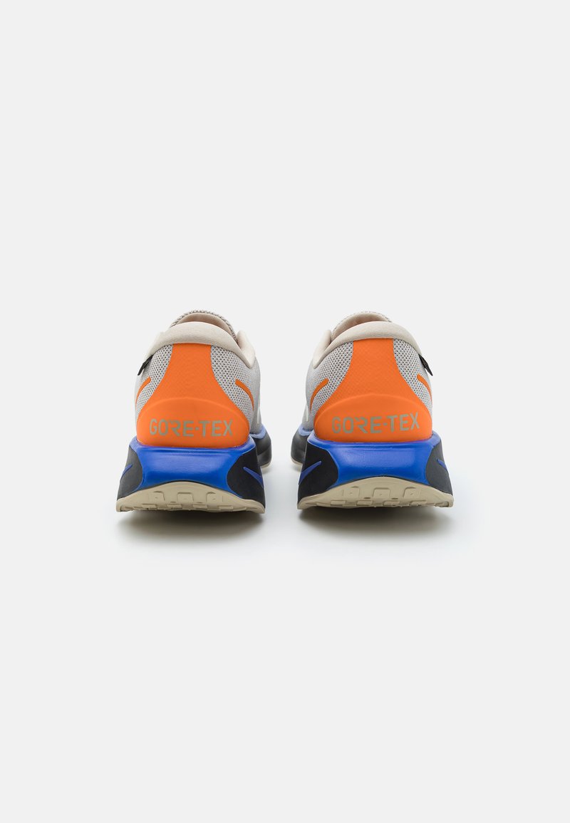 Zapatillas de running GORE-TEX con un upper texturizado en beige, acentos vibrantes en naranja, una mediasuela azul y una suela negra con un patrón resistente.