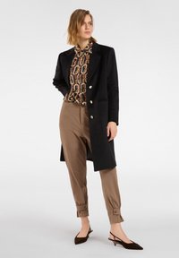 Frau trägt eine gemusterte Bluse, schwarzen langen Mantel mit Knöpfen, beige zulaufende Hose mit Umschlagdetails und dunkle spitze Slingback-Schuhe.