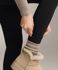 Bežna gležnjača z umetnim krznom, črne legice in rebraste bež nogavice, potegnjene čez črno tkanino. Roka prilagaja nogavico na gležnju.