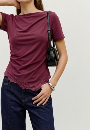 Modèle portant un haut bordeaux drapé avec une bordure en dentelle, un jean foncé, un sac à bandoulière noir et un bracelet en argent.