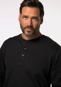 Schwarzes Langarm-Henley-Shirt aus geripptem Stoff mit Rundhalsausschnitt und drei Knöpfen in kontrastierendem Kupfer. 