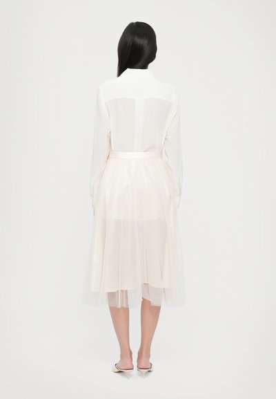 Transparante witte blouse met een kraag, gecombineerd met een ivoor kleuren tule rok. De rok heeft een gelaagd ontwerp, een strak middel en een vloeiende silhouet.