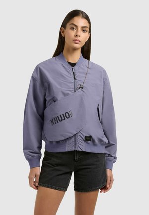 Young woman wearing a purple bomber jacket, black denim shorts, and a crossbody bag with "KHUJO 2001" text on a plain background.