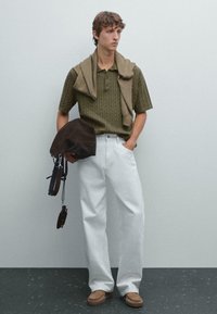 Un giovane uomo è in piedi contro un muro grigio chiaro, indossando un polo oliva, pantaloni bianchi e scarpe marroni, mentre tiene in mano una borsa marrone con una tracolla.