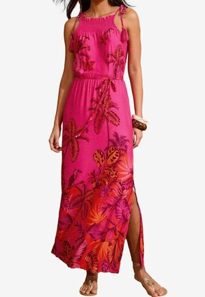 Kvinde iført en ærmeløs pink maxi kjole med rødt tropisk blad- og fugleprint, sidesplit, bindebånd i taljen og skulderkvaster, kombineret med sandaler.