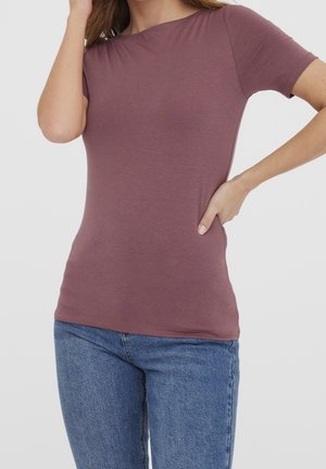 Femme portant un t-shirt moulant mauve à manches courtes et un jean bleu, posant avec une main sur la hanche et l'autre touchant ses cheveux.
