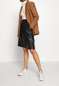 Brun kavaj, vit ribbad polotröja, svarta lädershorts med fåll och vita sneakers med pastellaccent. Ren, modern silhuett.