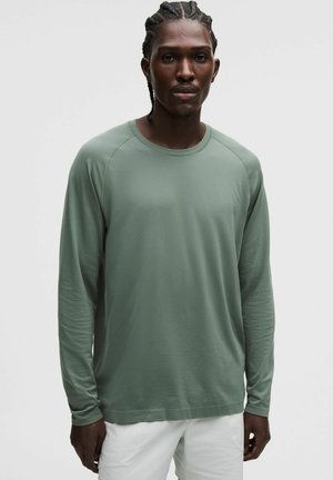 Mand med flettet hår iført en lysegrøn langærmet skjorte og hvide bukser, står foran en ensfarvet lys baggrund.