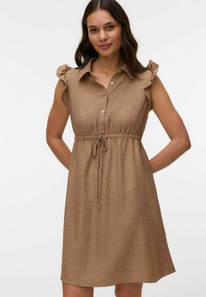 Žena s dlhými tmavými vlasmi, oblečená v bezrukávových svetlohnedých šatách po kolená s volánmi na ramenách a šnúrkou v páse, stojaca s rukami za chrbtom.