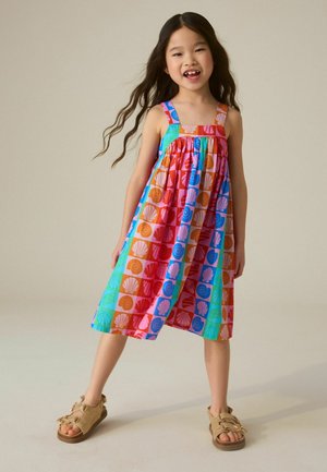 Ung pige med langt hår iført en farverig, ærmeløs kjole med skalmønstre og beige sandaler, stående med benene fra hinanden på en ensfarvet baggrund.