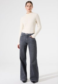 Lichtcrème gebreide trui met hoge hals, gecombineerd met donkergrijze wijde jeans met een standaard voorzak en een knoopsluiting.