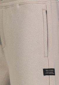 Beige Jogginghosen aus strukturiertem Stoff mit seitlichen Taschen und einem schwarzen Etikett mit weißem Text an der linken Hüfte.