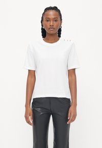 Hvid bomulds t-shirt med korte ærmer, rund hals og tre dekorative knapper på venstre skulder. Bæres med sorte læderbukser.
